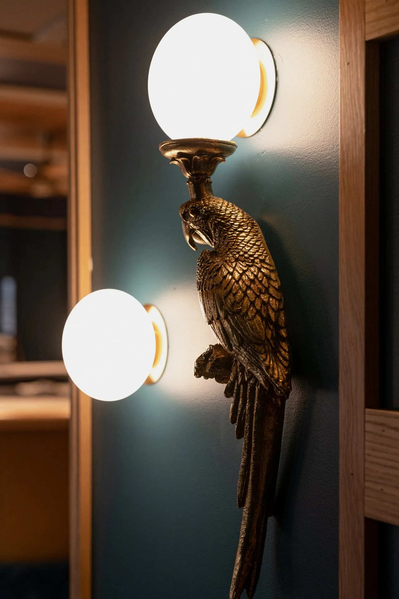 A gold-colored wall sconce in a bird shape holds a round white light bulb, mounted on a dark green wall.