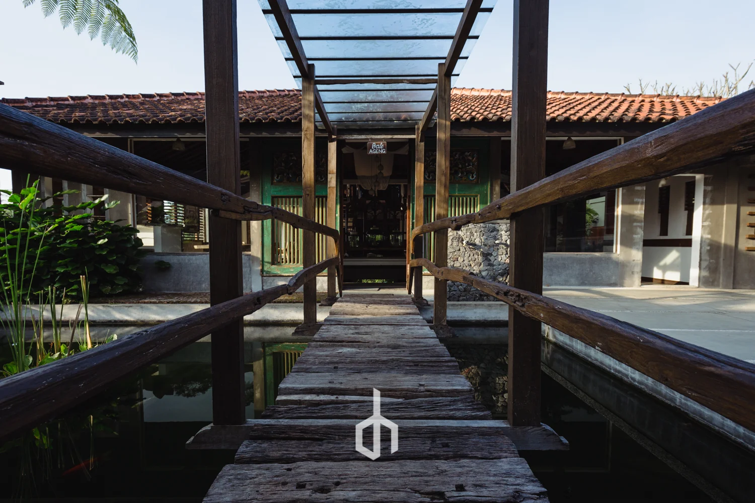 A wooden footbridge leading to a house with a tiled roof and a covered porch, surrounded by lush greenery and water.