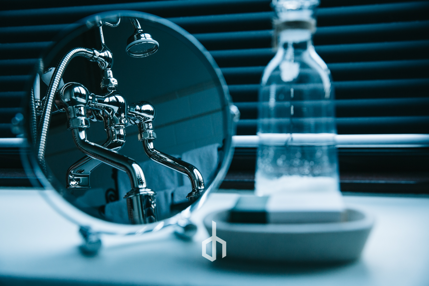 A small round mirror reflecting a chrome sink faucet with a showerhead attachment. A glass bottle is on a dish nearby, with a dark background of horizontal blinds.