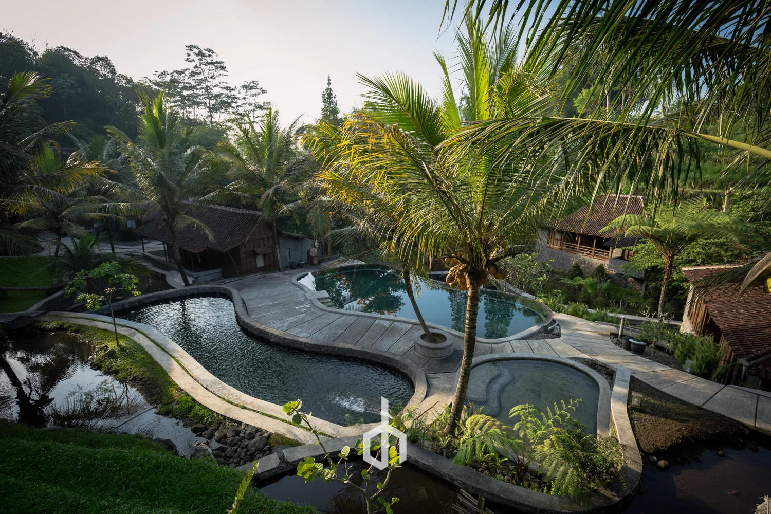 Resort with multiple pools surrounded by tropical plants and trees, with traditional-style buildings in the background.