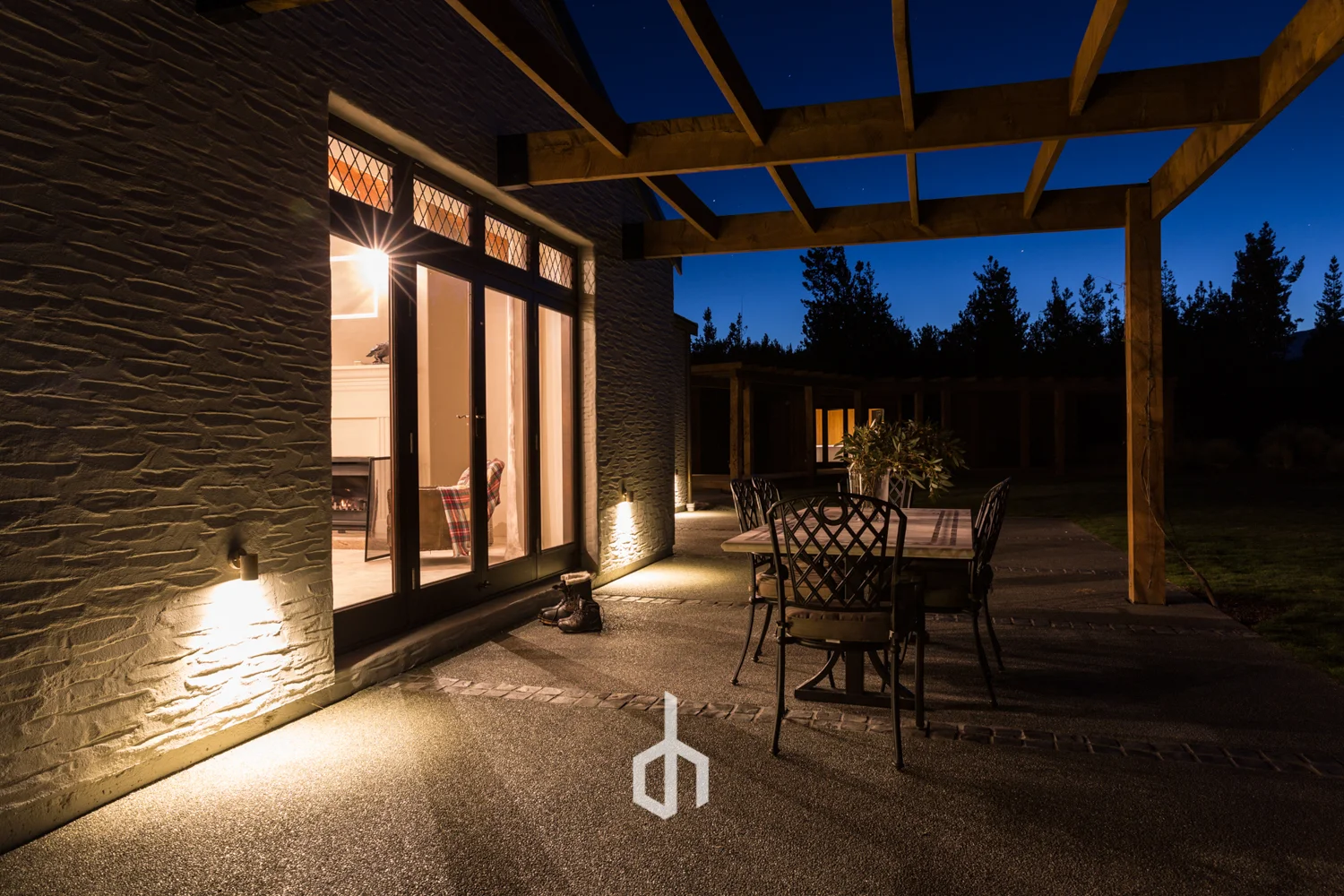 Nighttime view of a house's outdoor patio area with a table and six chairs, illuminated by exterior lights, with a view of trees and a dark sky in the background.