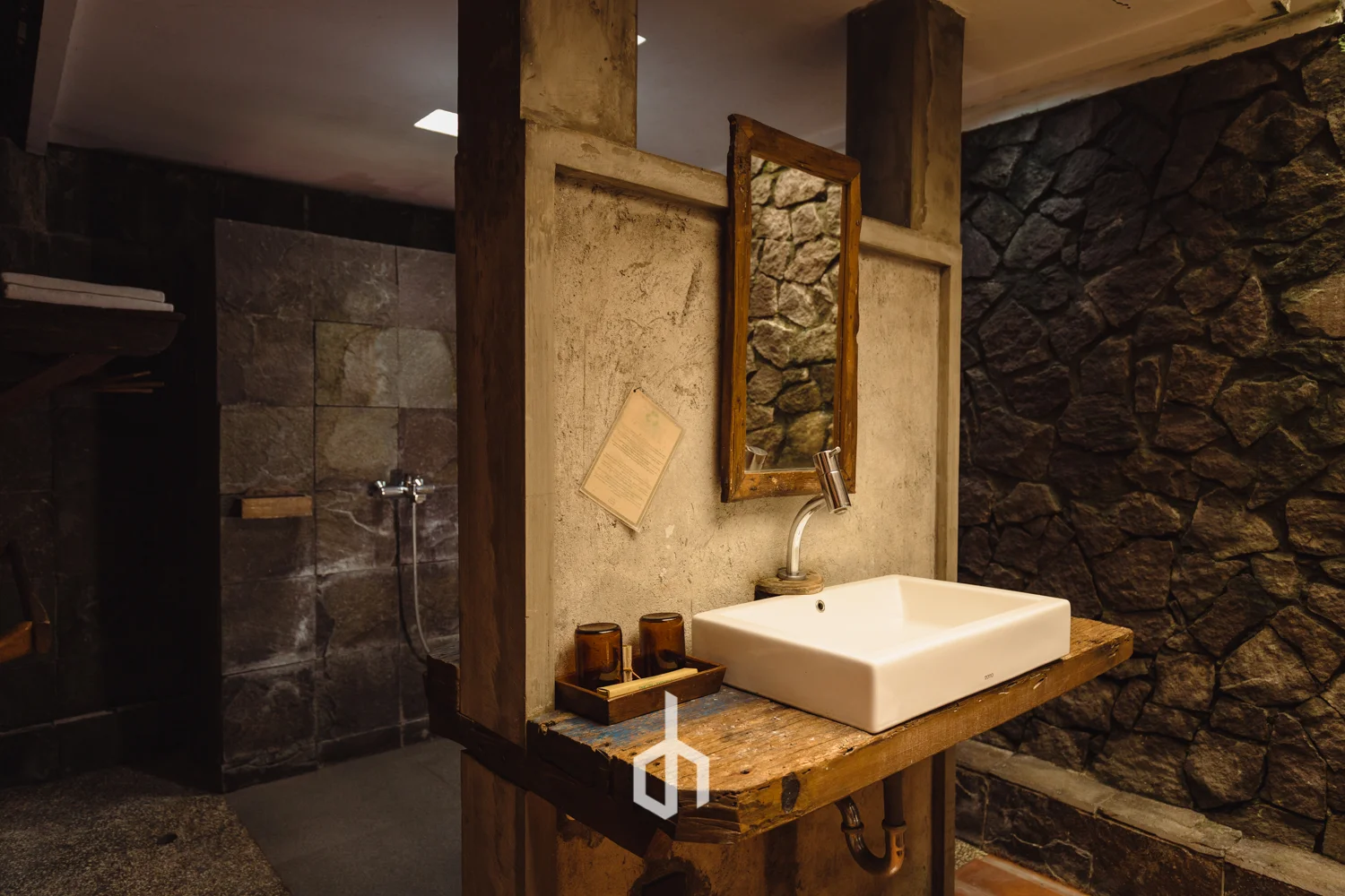 Rustic bathroom sink with a square white basin on a wooden countertop, a small wooden tray with glasses, a mirror with a wooden frame, stone walls, and a section of a shower area visible in the background.