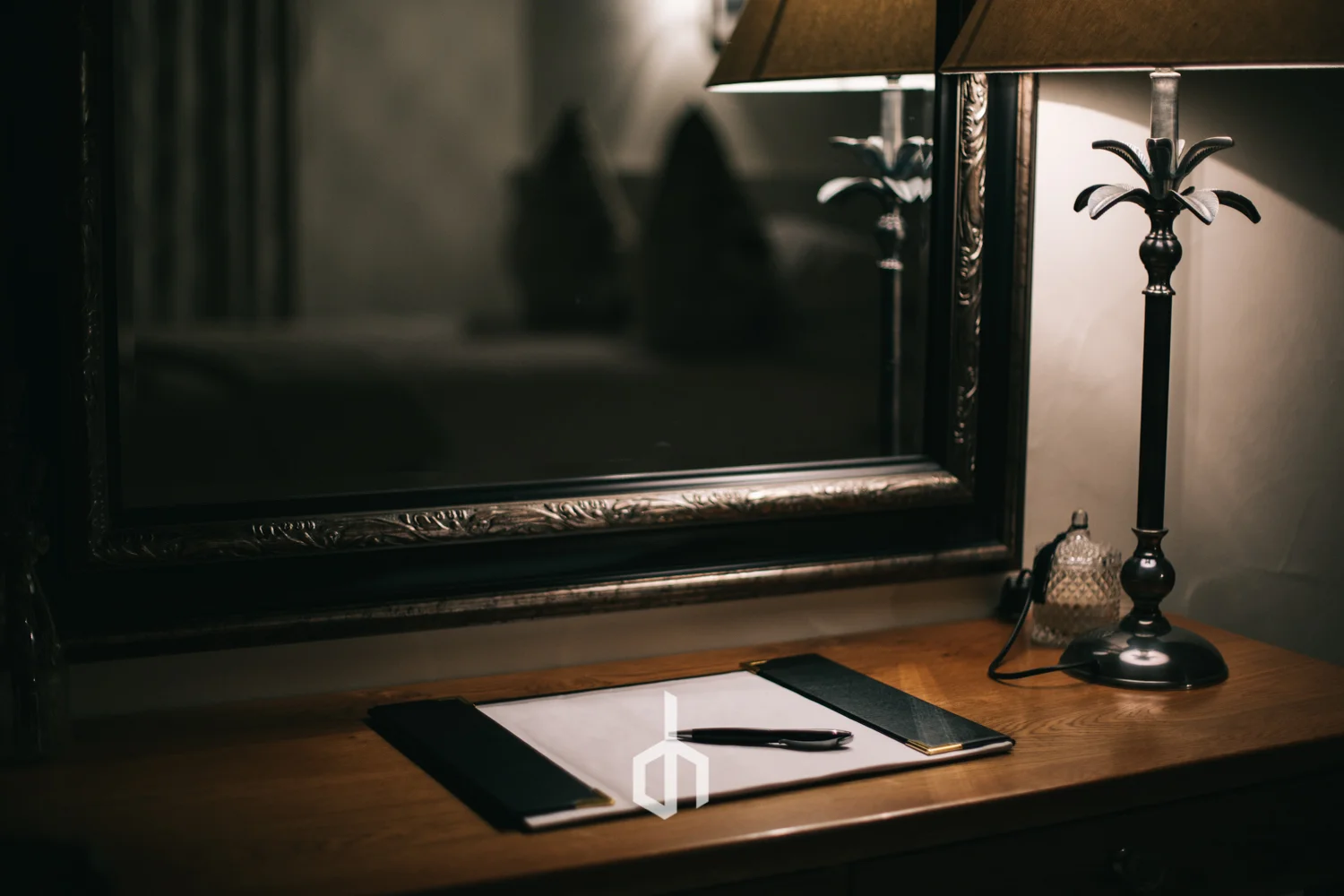 A wooden desk with a notebook, pen, and a ruler, illuminated by a table lamp beside a mirror.