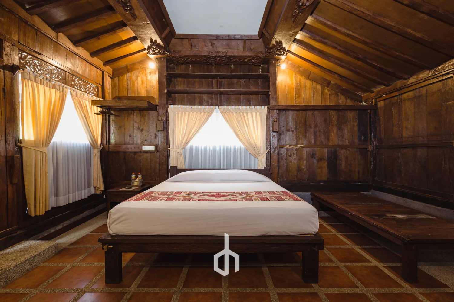 A cozy bedroom with wooden walls, a bed with white bedding and a decorative runner, beige curtains on windows, and warm lighting.