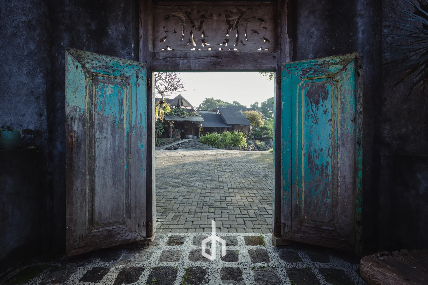 Open wooden door with weathered turquoise paint, leading to a courtyard with stone paving and traditional houses in the background, with lush greenery and sunlight.