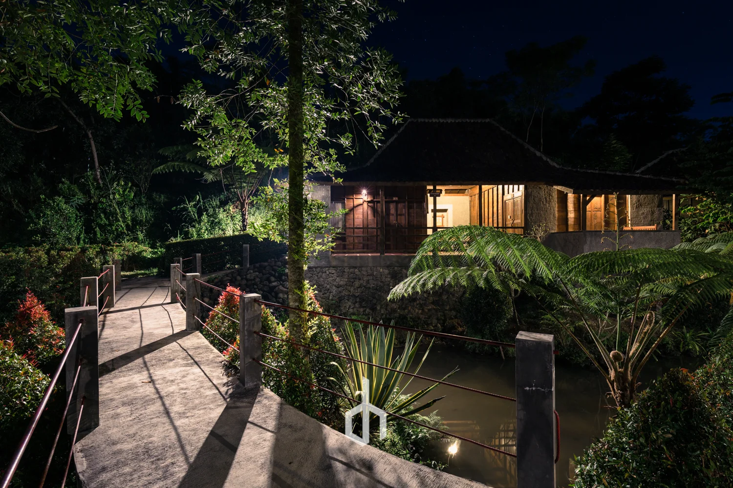 A illuminated traditional wooden house with a thatched roof located next to a small waterway at night, with a gravel pathway and lush greenery around.