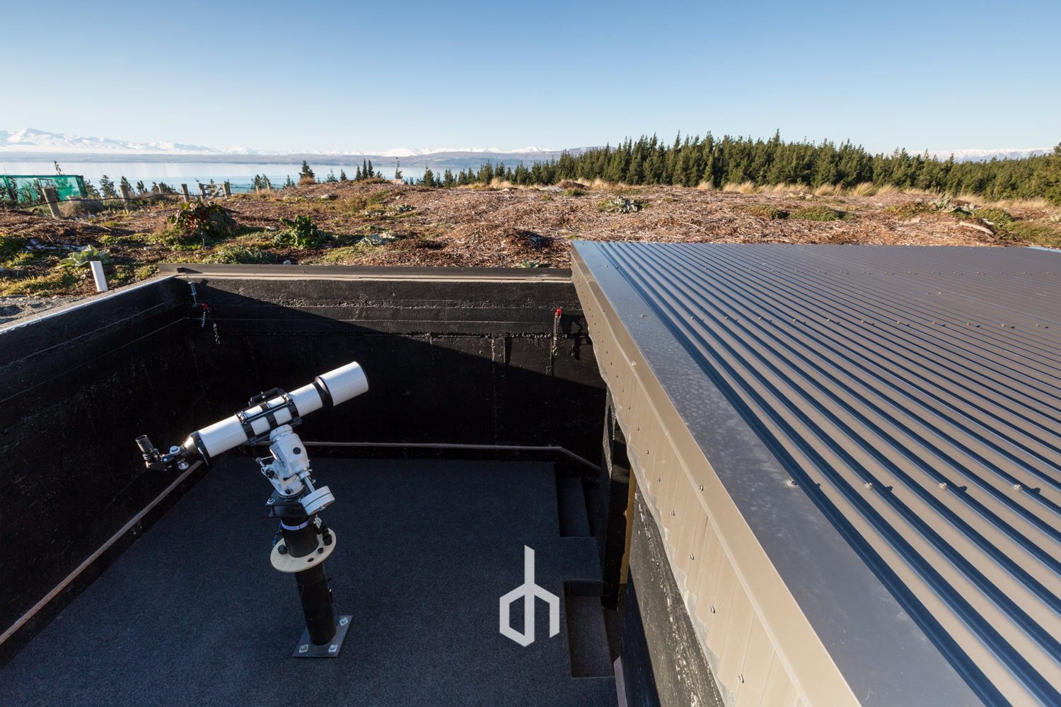 A telescope with a white and black body mounted on a stand on a flat rooftop, with a corrugated metal roof extending to the right. The rooftop is surrounded by black material, with a scenic landscape of trees, brown soil, and mountains in the backgro