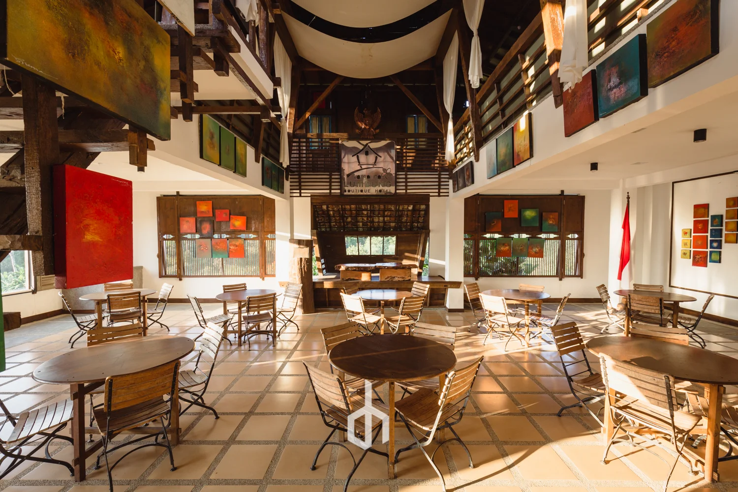 Interior of a cafe or restaurant with wooden tables and chairs, colorful abstract paintings on the walls, and natural sunlight streaming through the windows.