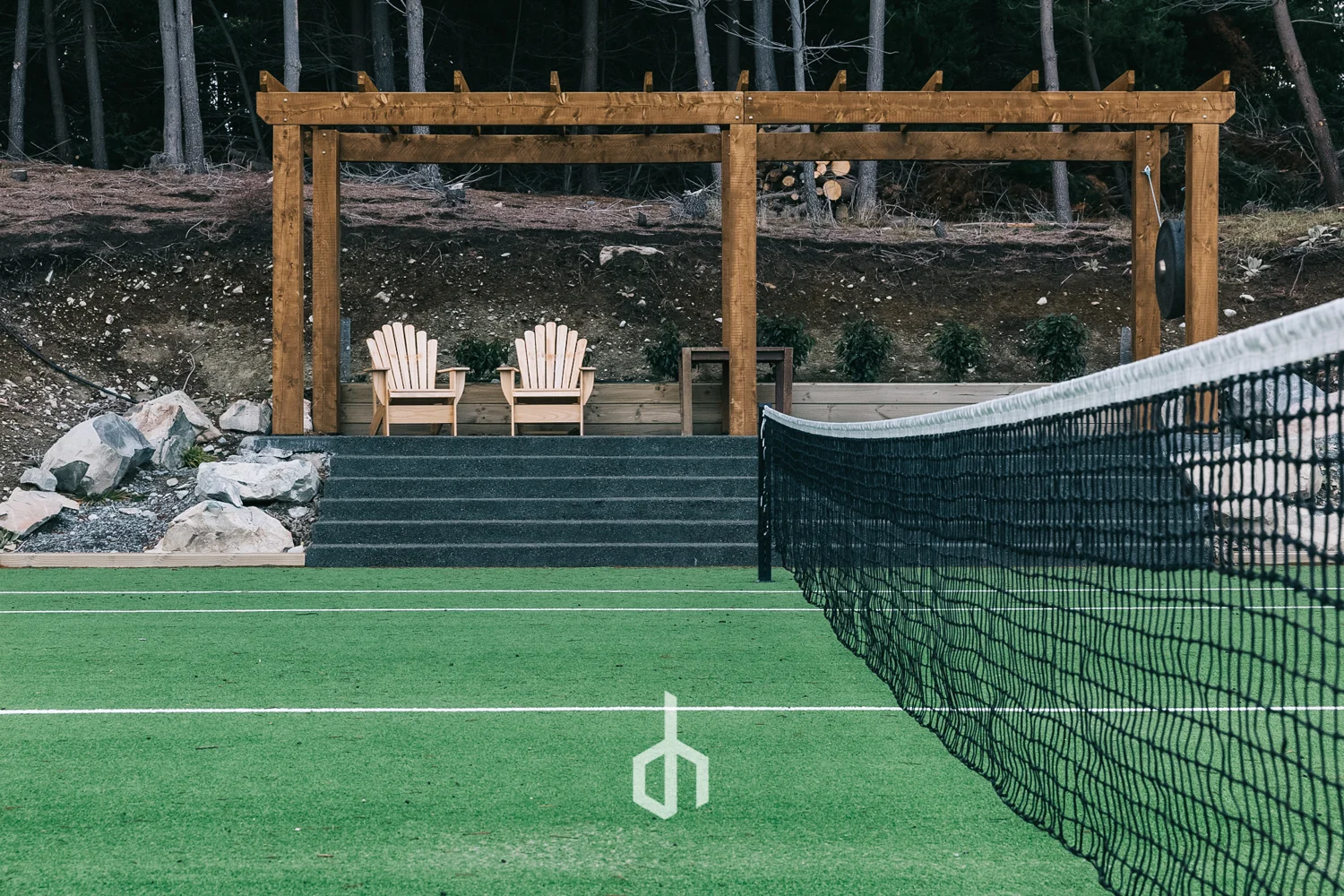 A tennis court with green surface and white lines, black net, concrete stairs leading to a wooden seating area with two Adirondack chairs, and a wooded hillside in the background.