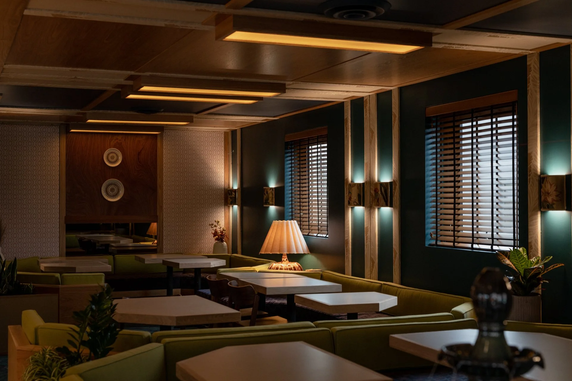 A cozy interior lounge with green seating, wooden wall accents, and windows with wooden blinds, illuminated by soft lighting and a table lamp.