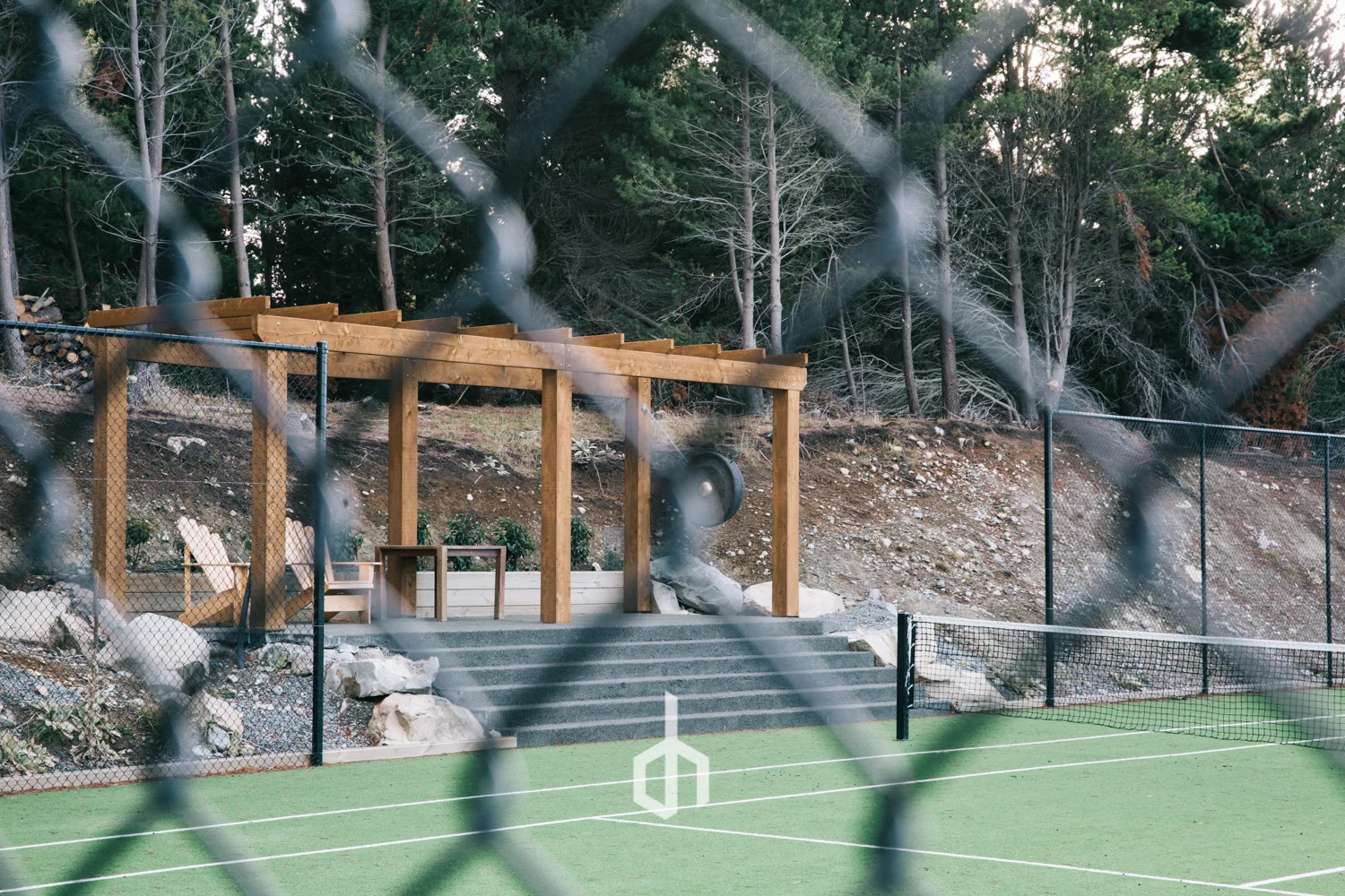 A tennis court with a green surface, a black net, and a wooden structure with a slide behind a chain-link fence, with trees in the background.