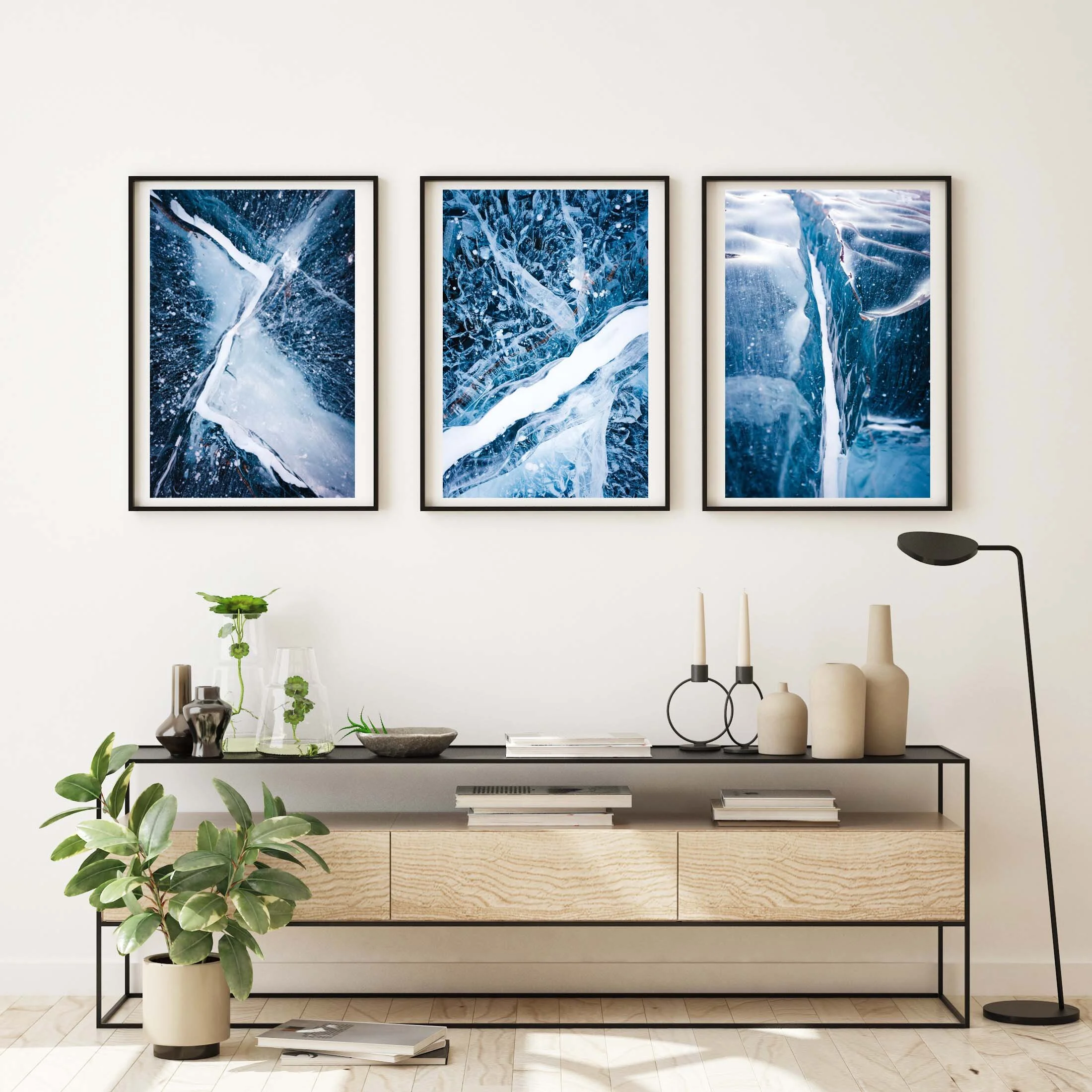 A minimalist interior space with three large framed blue abstract art pieces on a white wall, a black metal console table with various decorative objects including plants, vases, and candles, and a wooden cabinet below holding books, with a green potted plant on the floor in front.