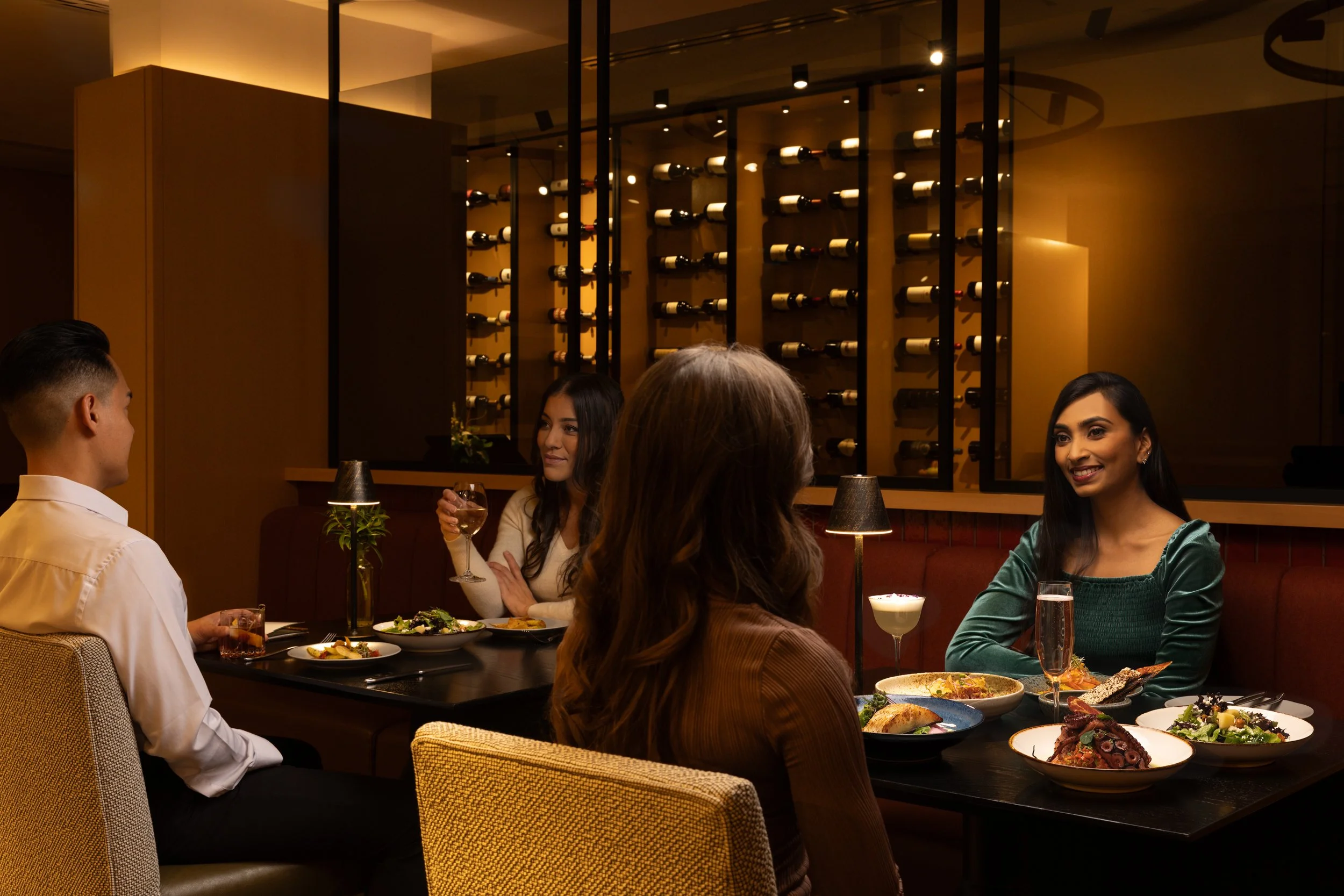 Group of five people enjoying dinner in a wine-themed restaurant with a wine rack on the wall in the background.