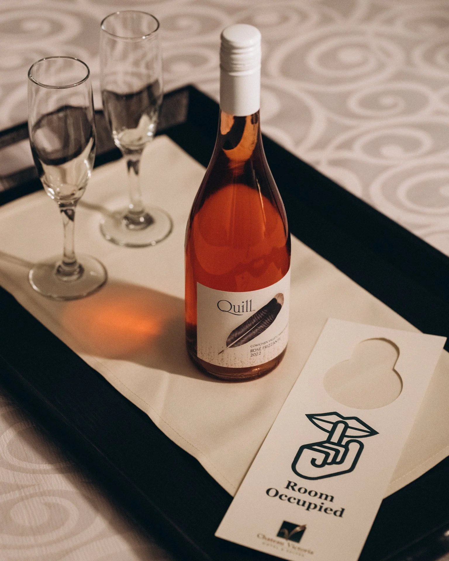 A bottle of rosé wine labeled 'Quill' on a table with two empty champagne flutes and a white paper with a black outline of lips and the text 'Room Occupied'.