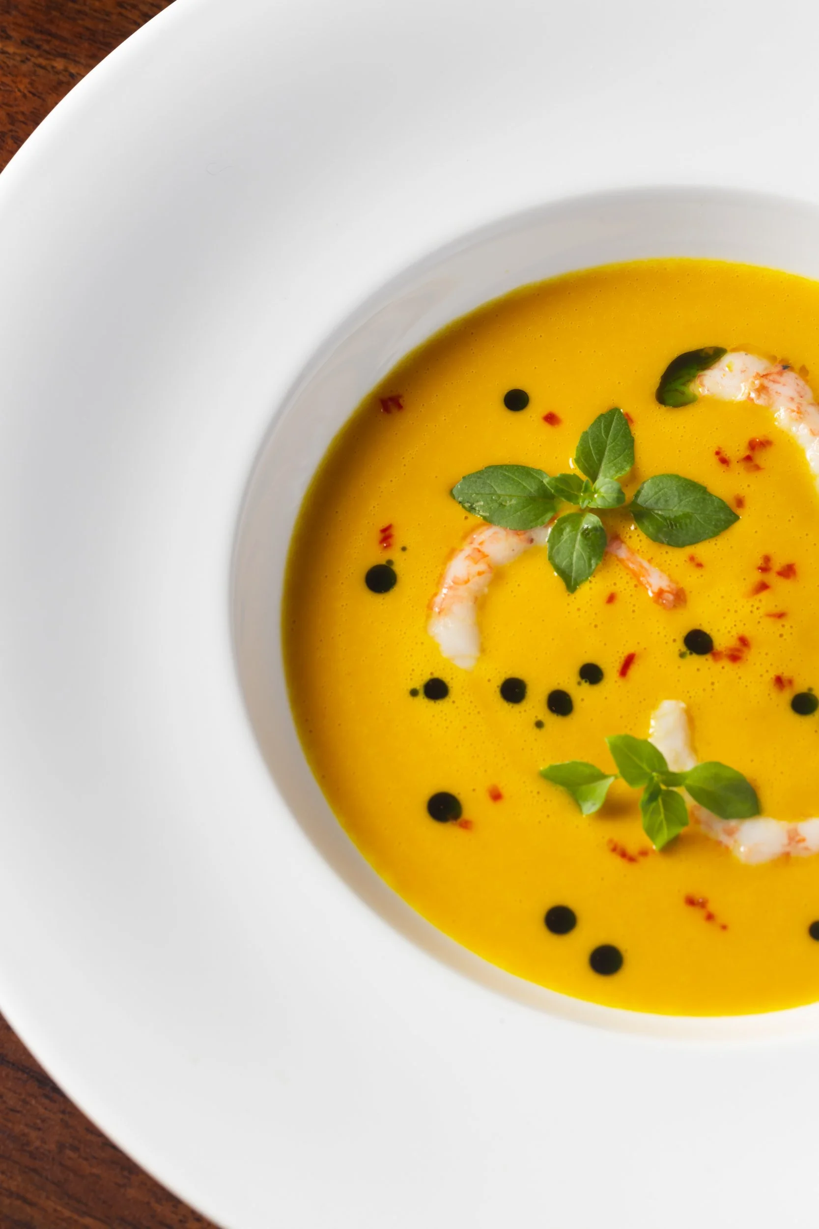 A bowl of bright yellow soup garnished with fresh green herbs, black pepper, and small pieces of seafood or shrimp, on a wooden surface.