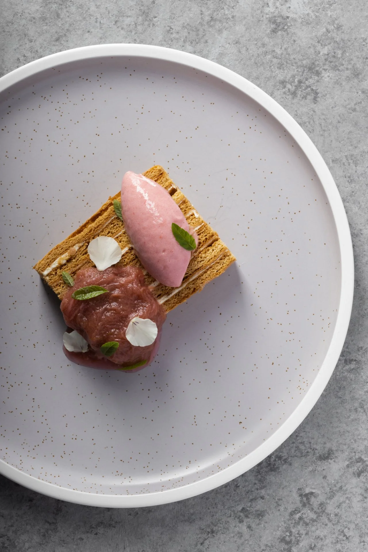 A plate with layered cake topped with pink ice cream, garnished with edible flowers and green leaves, on a gray surface.