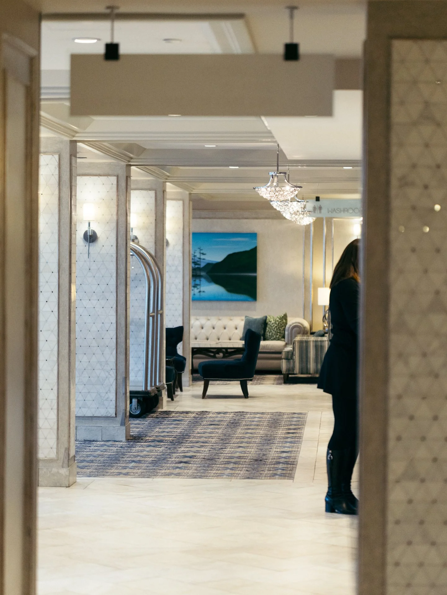 Inside a hotel lobby with cream-colored walls, chandeliers, a painting of a lake and mountains, sofas, chairs, and a person in dark clothing standing near a wall.