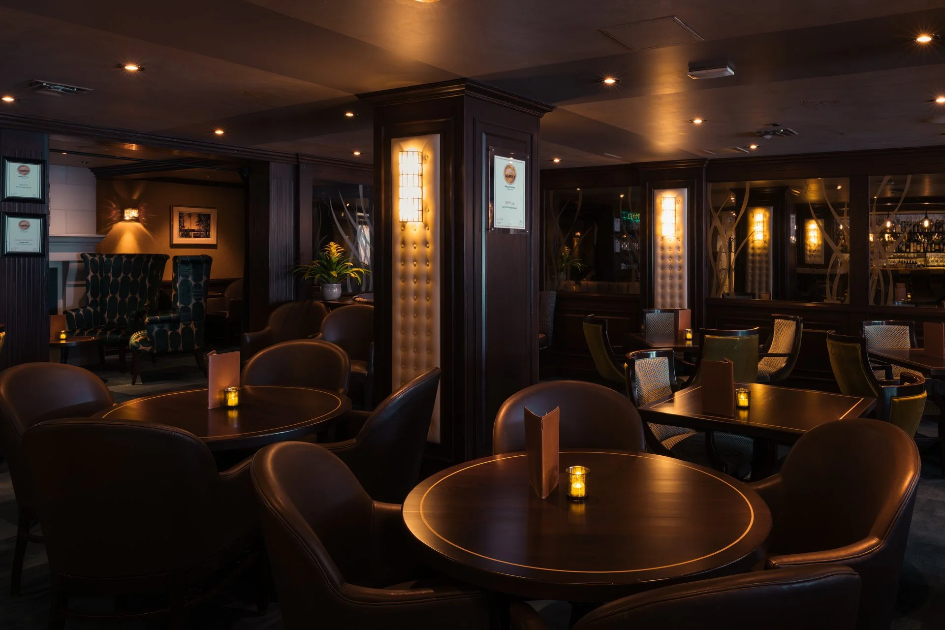 Interior of a dimly lit bar or lounge with dark wooden furniture, round tables with small candles, leather and patterned chairs, decorative wall panels with lighting, and a bar area in the background.