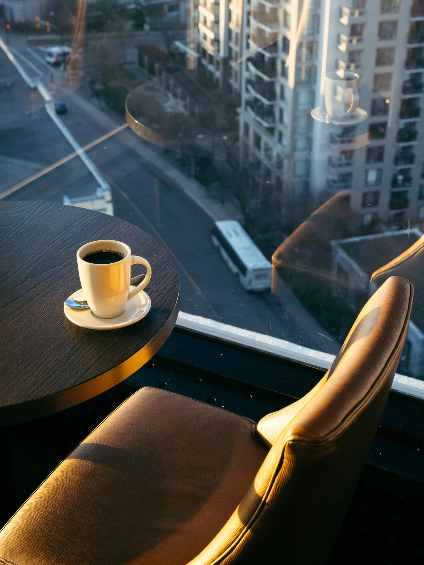 A white coffee cup filled with black coffee on a saucer with a spoon, placed on a dark, round table near a large window with a cityscape view including tall buildings and a bus.