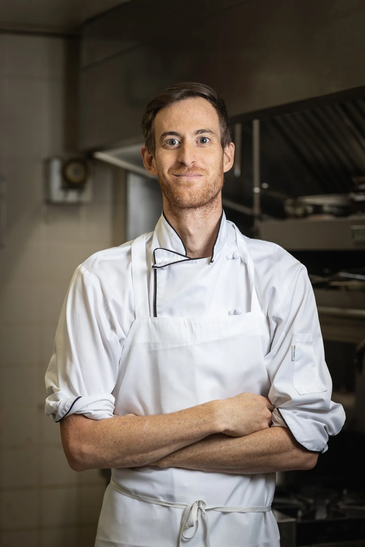 A male chef standing in a professional kitchen, wearing a white chef's coat and apron, with arms crossed and smiling at the camera.