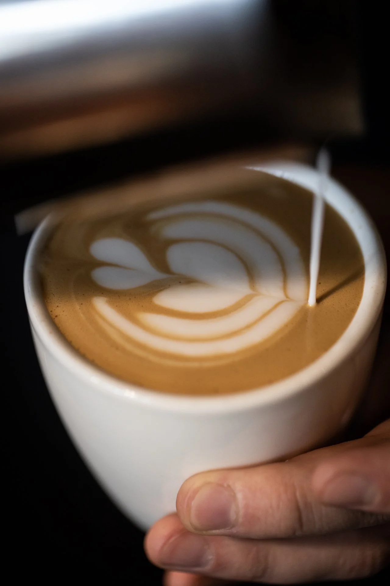 Close-up of a hand holding a white coffee cup with latte art in the shape of a leaf design, as hot coffee is being poured into it.