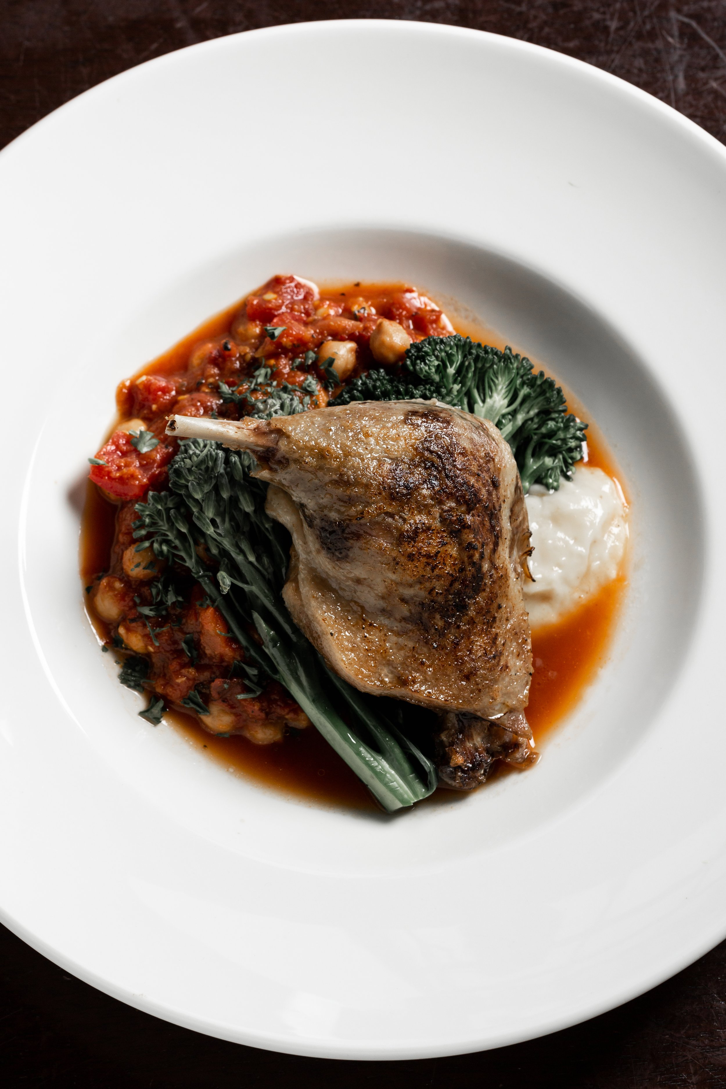 A white plate holding a cooked chicken thigh with crispy skin, garnished with cooked kale and served with a side of tomato-based stew and creamy mashed potatoes.