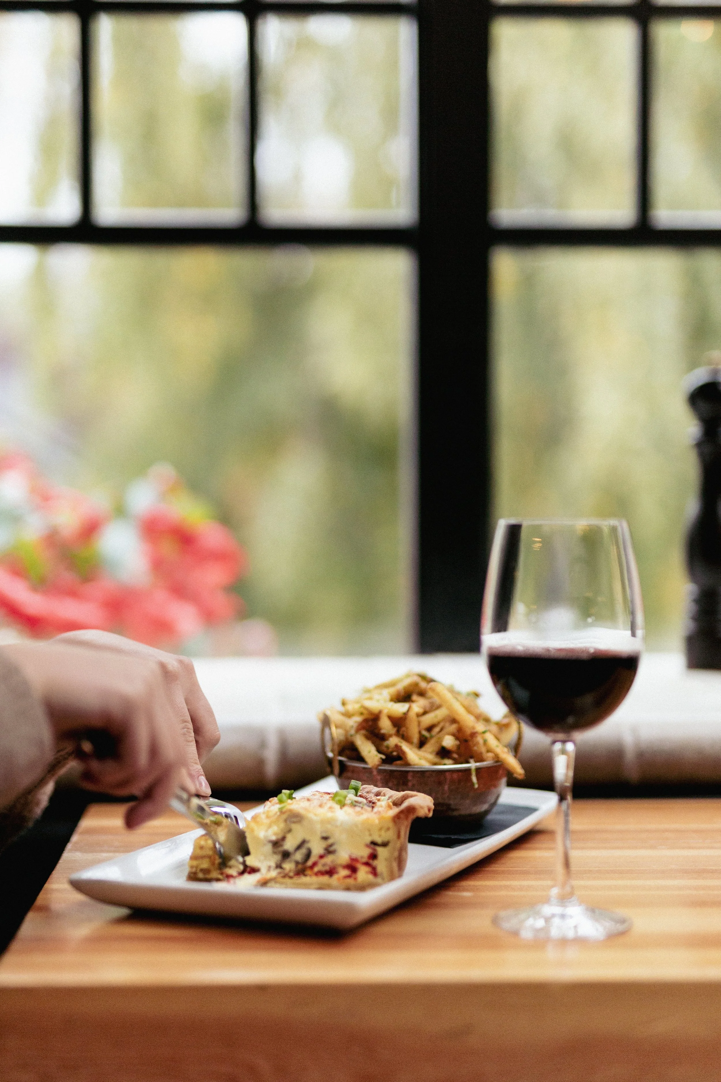 A dining scene with a glass of red wine, a plate of pasta, and a slice of quiche on a white plate on a wooden table near a large window with an outside view of trees.