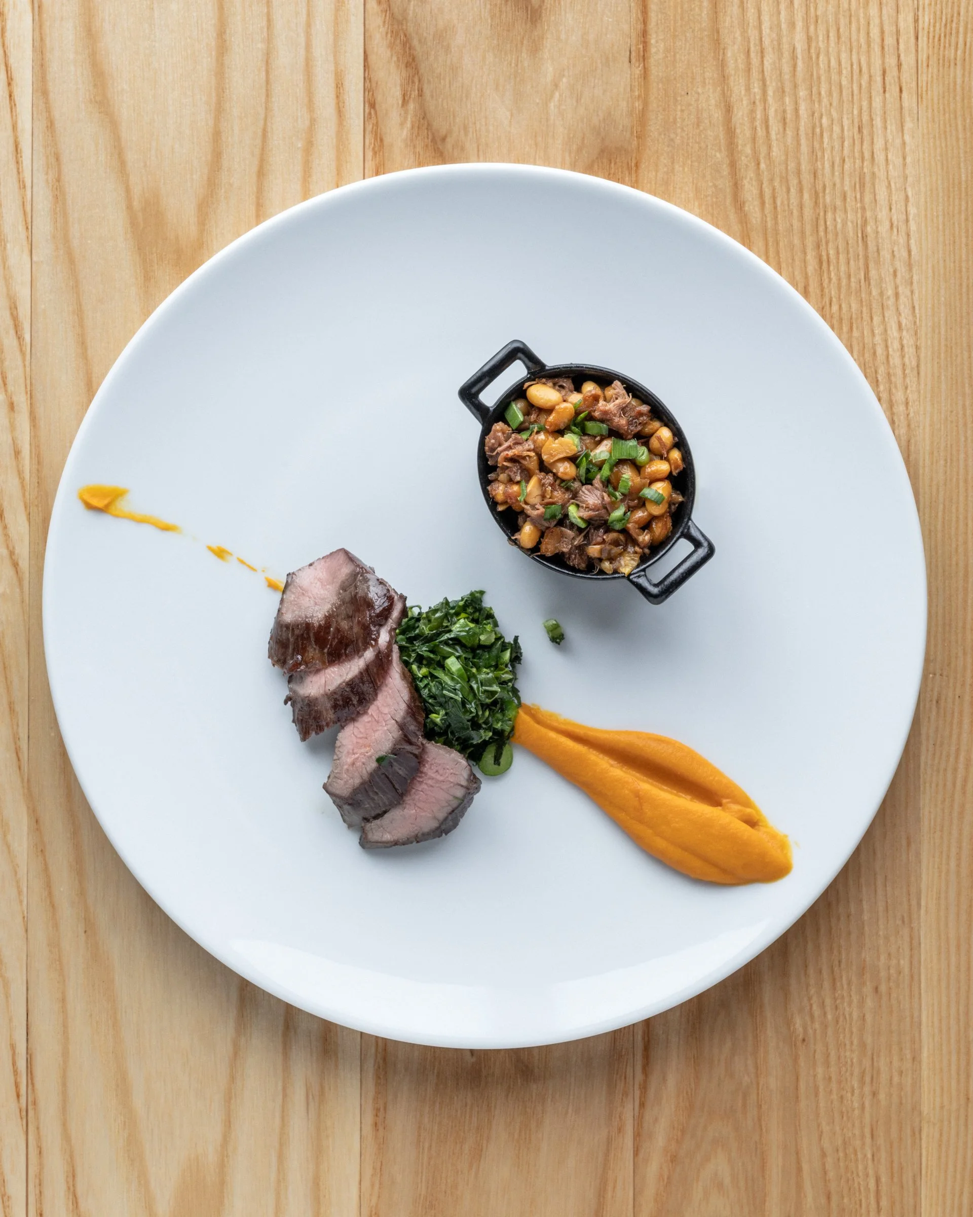 A white round plate on a wooden surface with slices of cooked meat, a dollop of yellow sauce, a small black bowl of beans with chopped green onions, and some chopped greens with a yellow sauce streak.