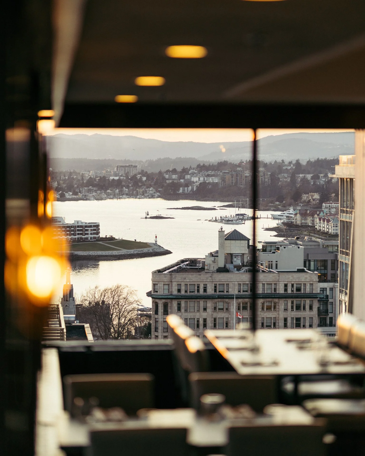 View of a river or bay with boats, surrounded by buildings and hills in the distance, seen through a window with reflections and blurred lights.