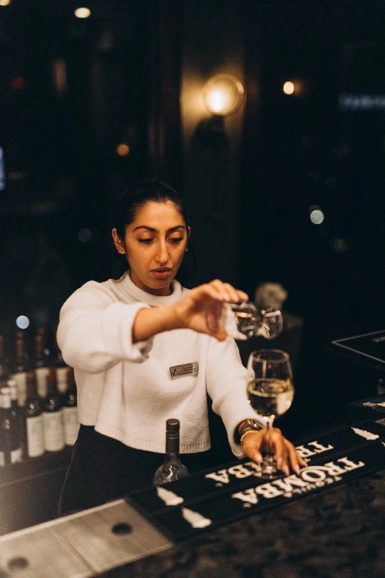 A woman with dark hair tied back, wearing a white sweater and a name badge, pours white wine into a glass at a bar counter in a dimly lit setting.