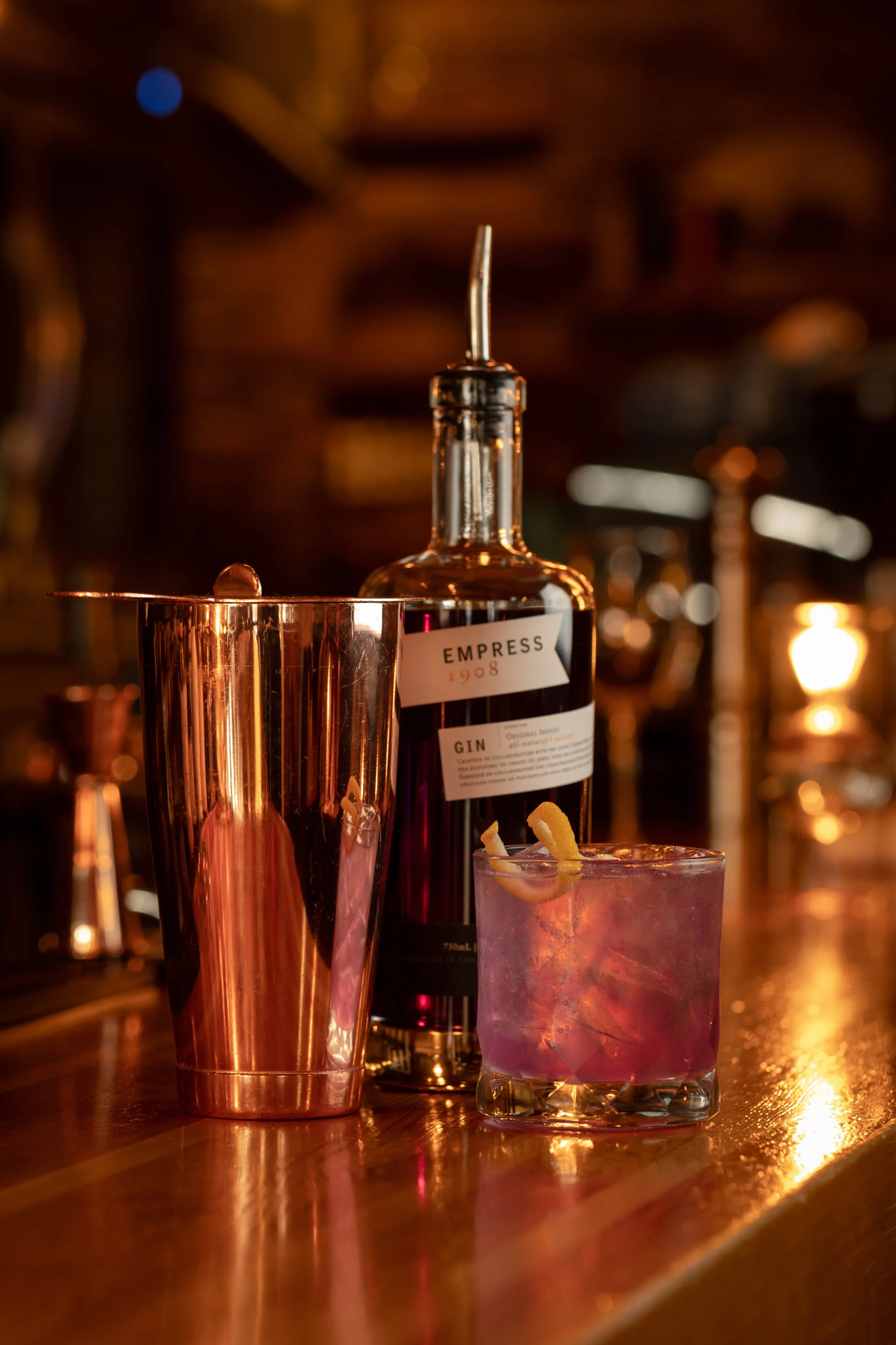 Close-up of a bar scene with a bottle of Empress gin, a pink cocktail garnished with lemon peel, and a reflective metallic cup on a wooden bar counter. The background features a warm, dimly lit ambiance.