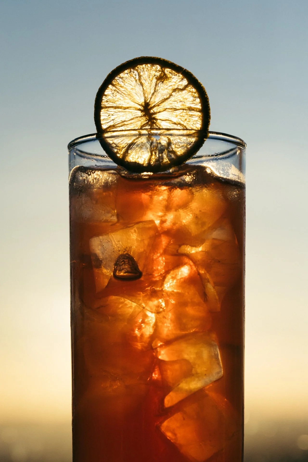 A tall glass filled with ice cubes and a dark beverage, garnished with a slice of lemon on the rim, with sunlight shining through the drink.