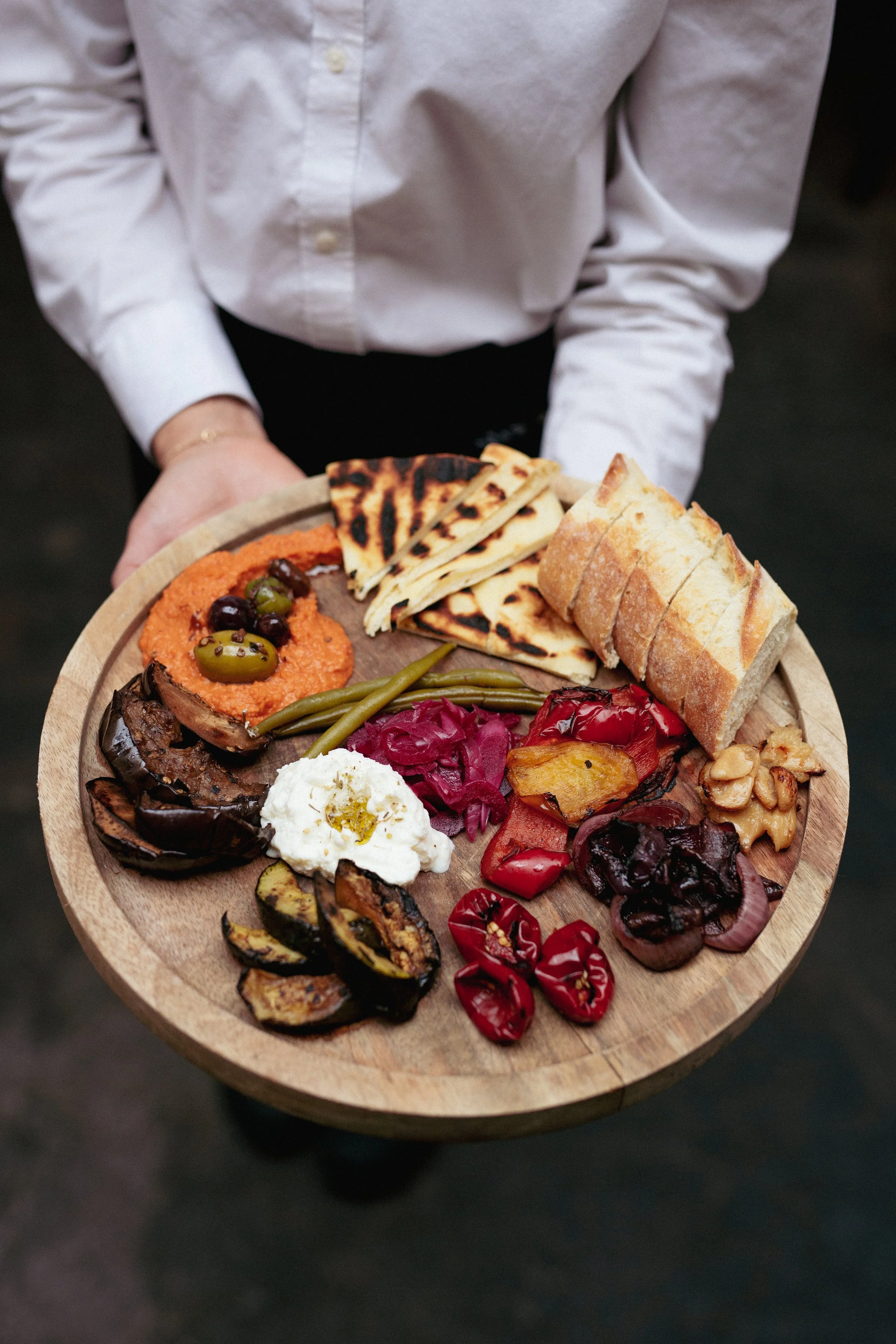 Person holding a round wooden platter filled with various Mediterranean appetizers, including grilled vegetables, cheese, pita bread, spreads, and pickled vegetables.
