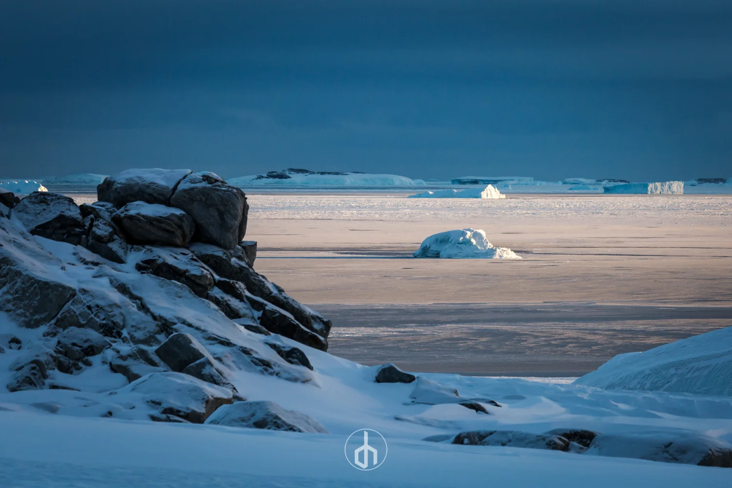 A snowy Arctic landscape with scattered icebergs and rocky shoreline under a cloudy sky.