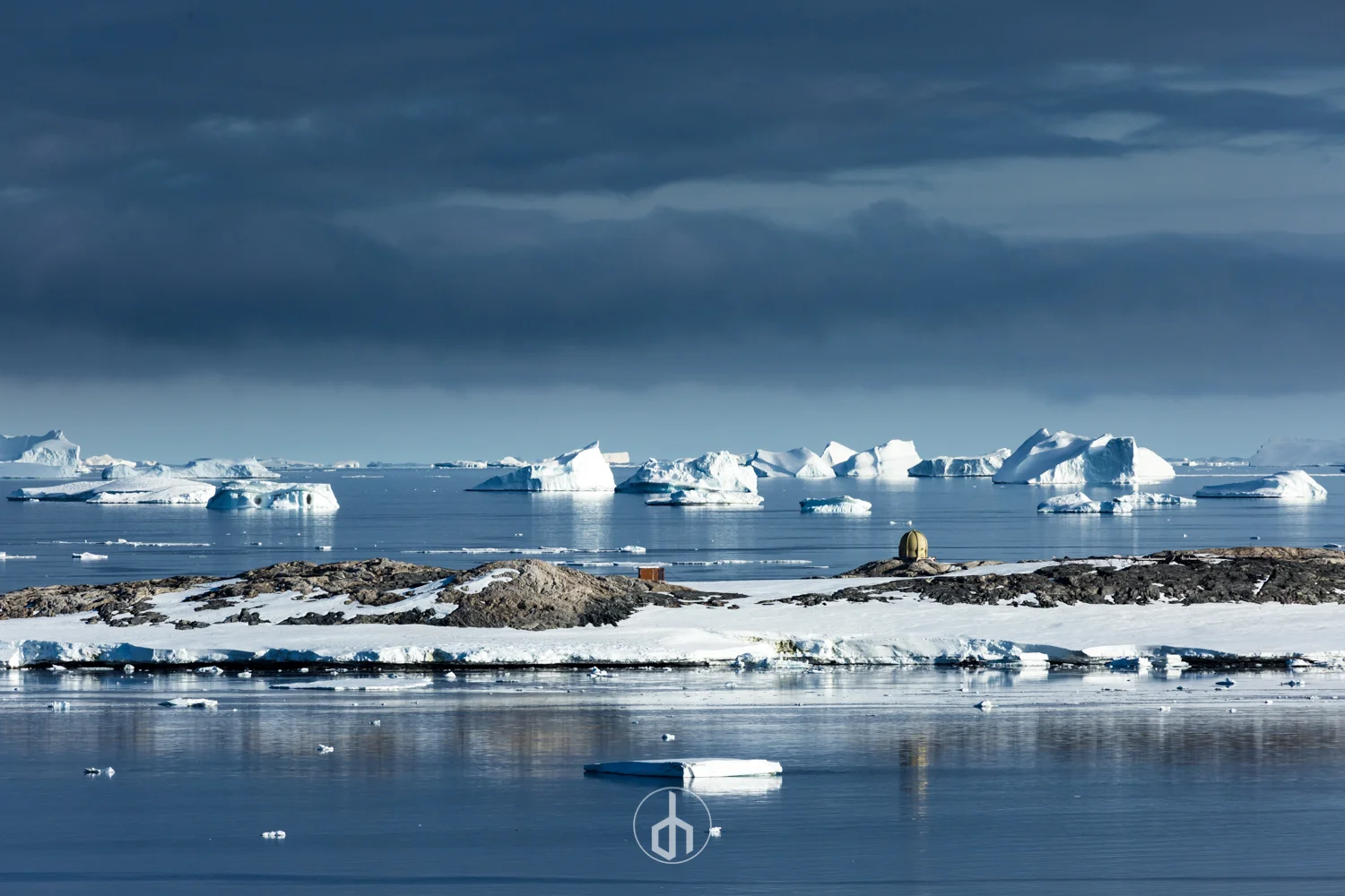 A cold, icy landscape showing a body of water with floating icebergs, a rocky shoreline with patches of snow, and a cloudy sky overhead.