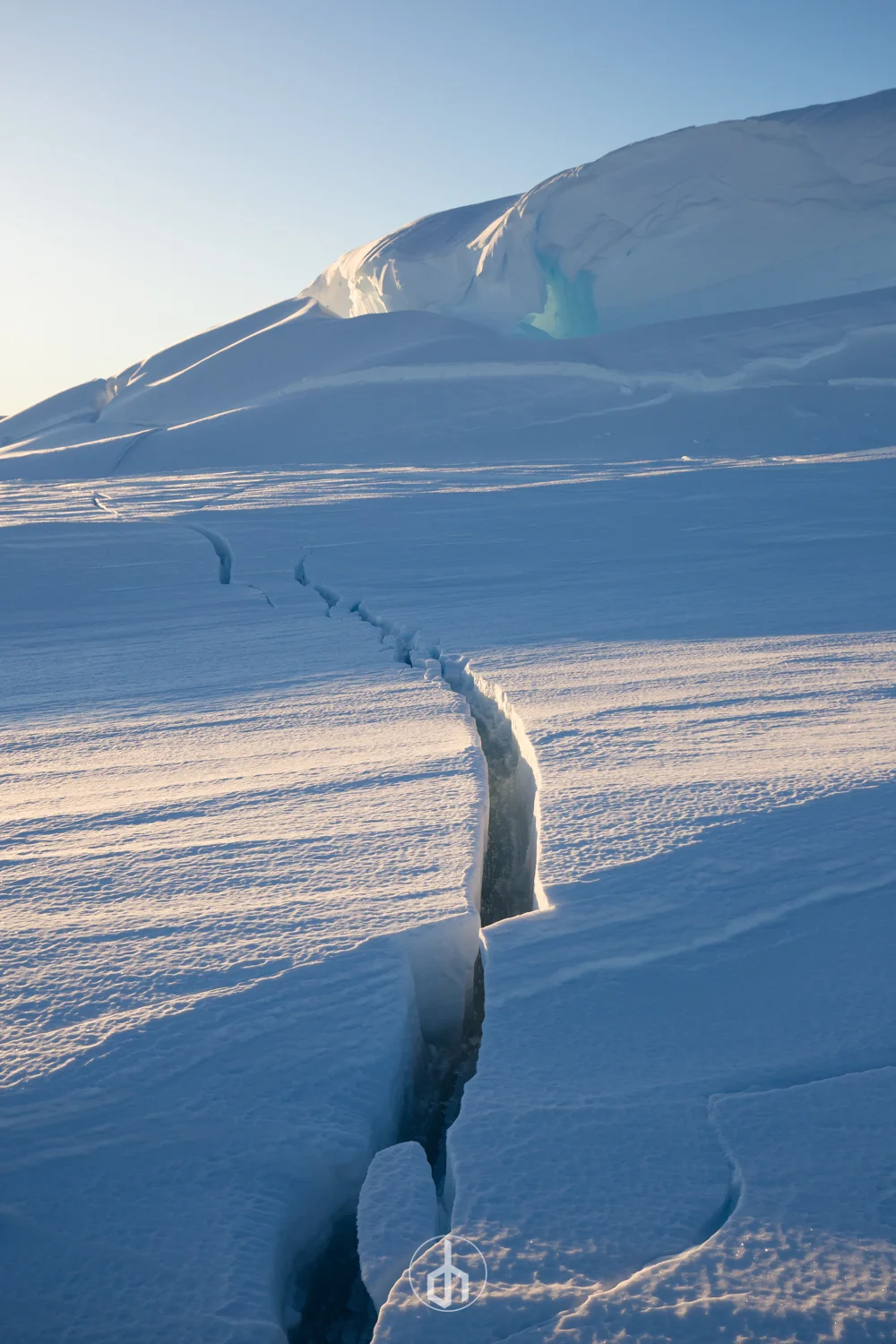 A snowy landscape with a large crack in the ice leading towards a snow-covered mountain under a clear sky.