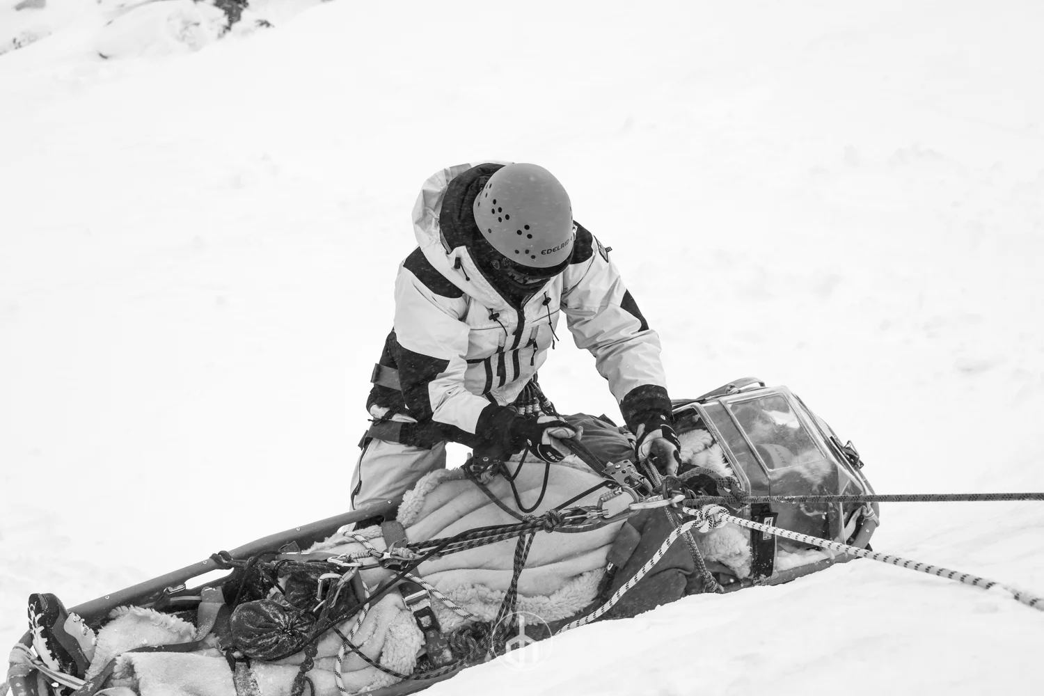 A person wearing winter gear, including a helmet and gloves, is kneeling next to a snow-covered rescue sled with equipment and a figure lying on it, holding onto ropes in a snowy environment.