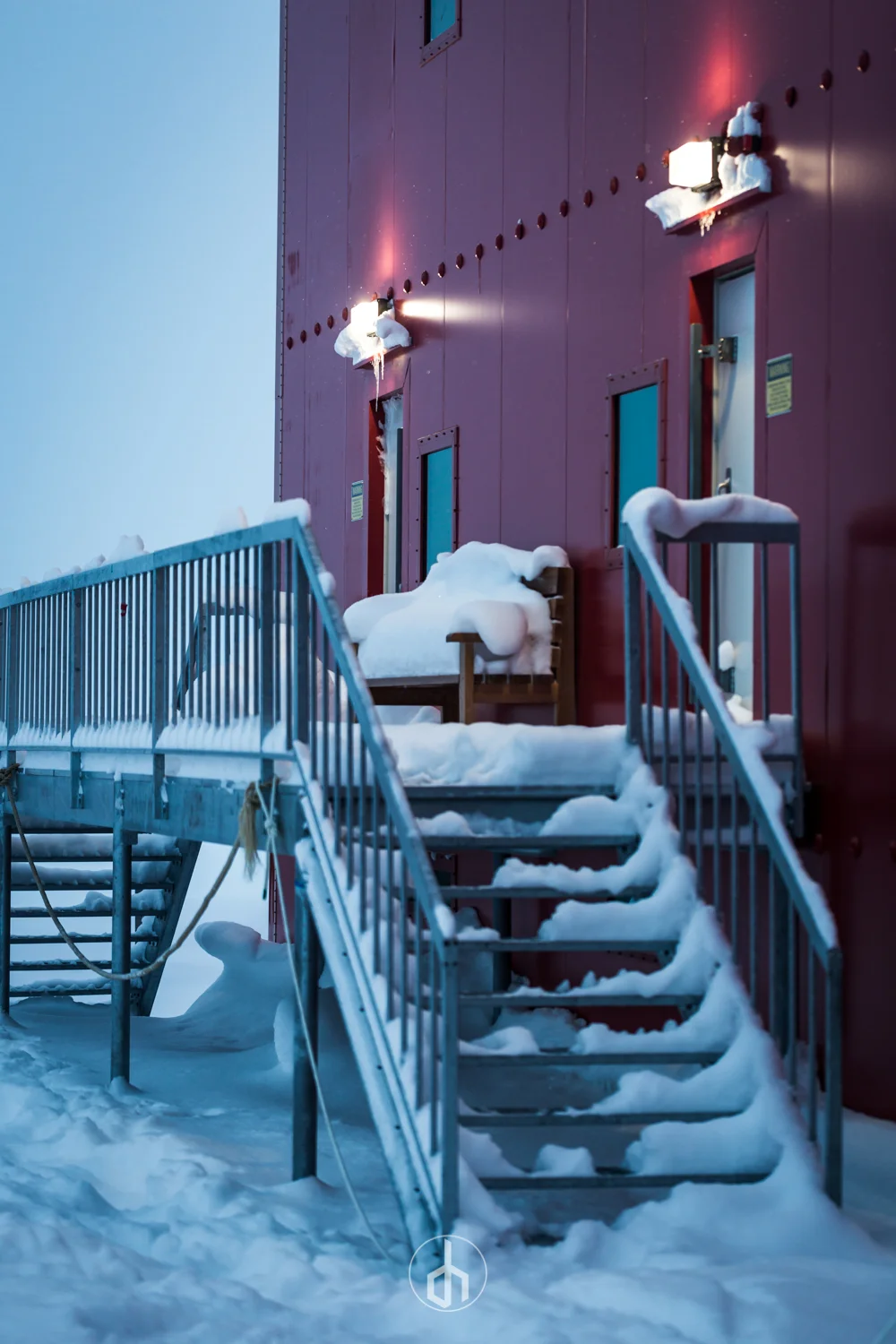 Snow-covered metal stairs leading to a red building with small windows and doors, snow piled on the railing and stairs, and snow-covered light fixtures mounted on the building's exterior.