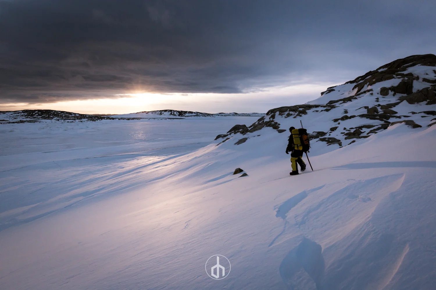 Person snowshoeing in a snowy, mountainous landscape at sunset or sunrise with dark clouds overhead.