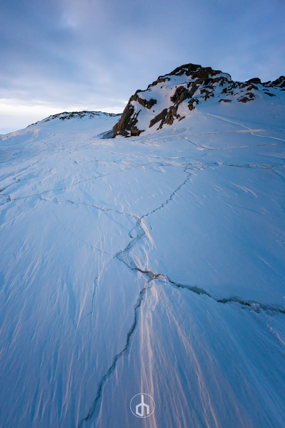 Snow-covered mountain landscape with visible cracks in the ice and a rocky peak in the distance under a partly cloudy sky.