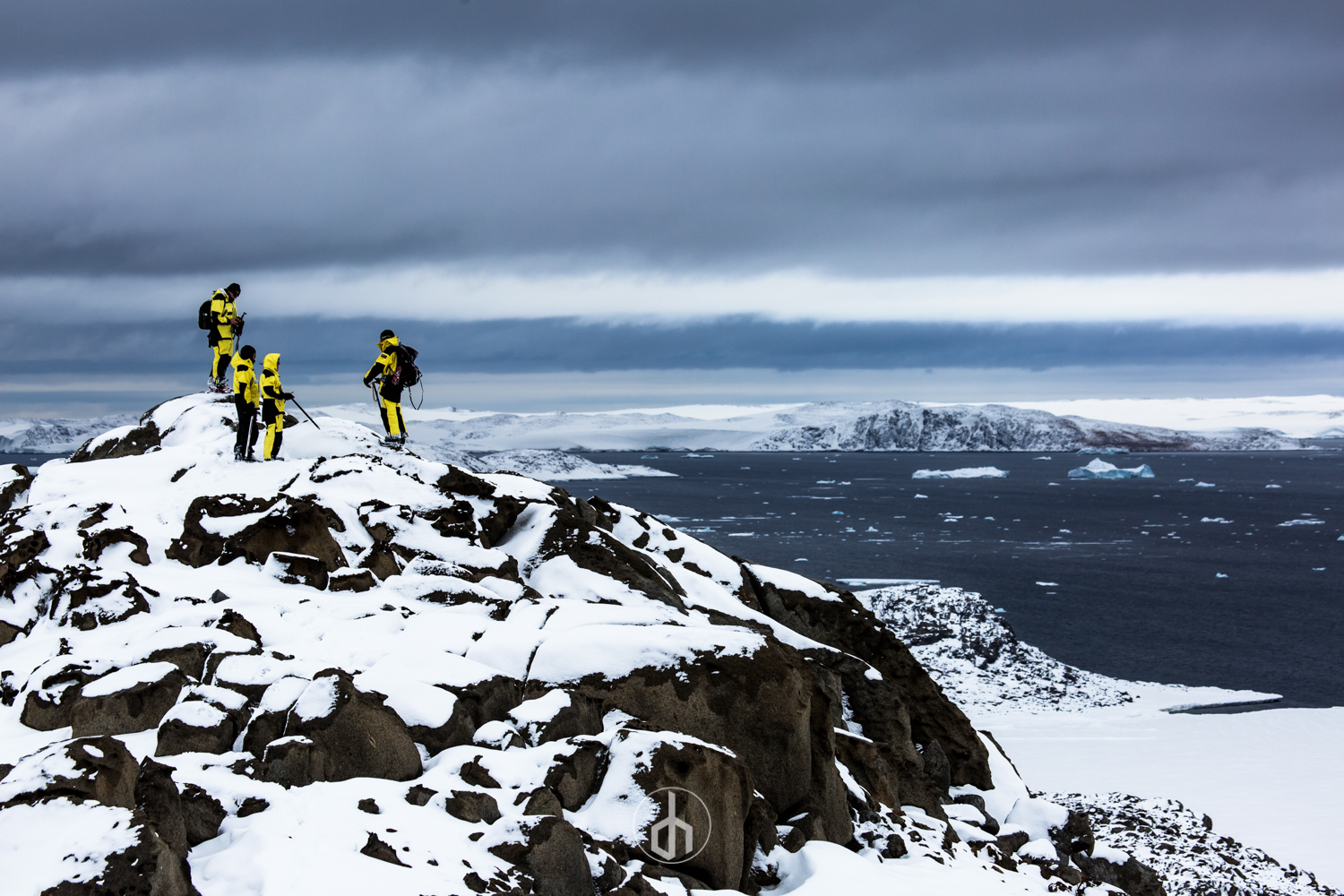 Group of five people in yellow and black winter clothing with backpacks, standing on snow-covered rocks on a cold, icy landscape with a snow-covered mountain and a body of water with icebergs in the distance under a cloudy sky.