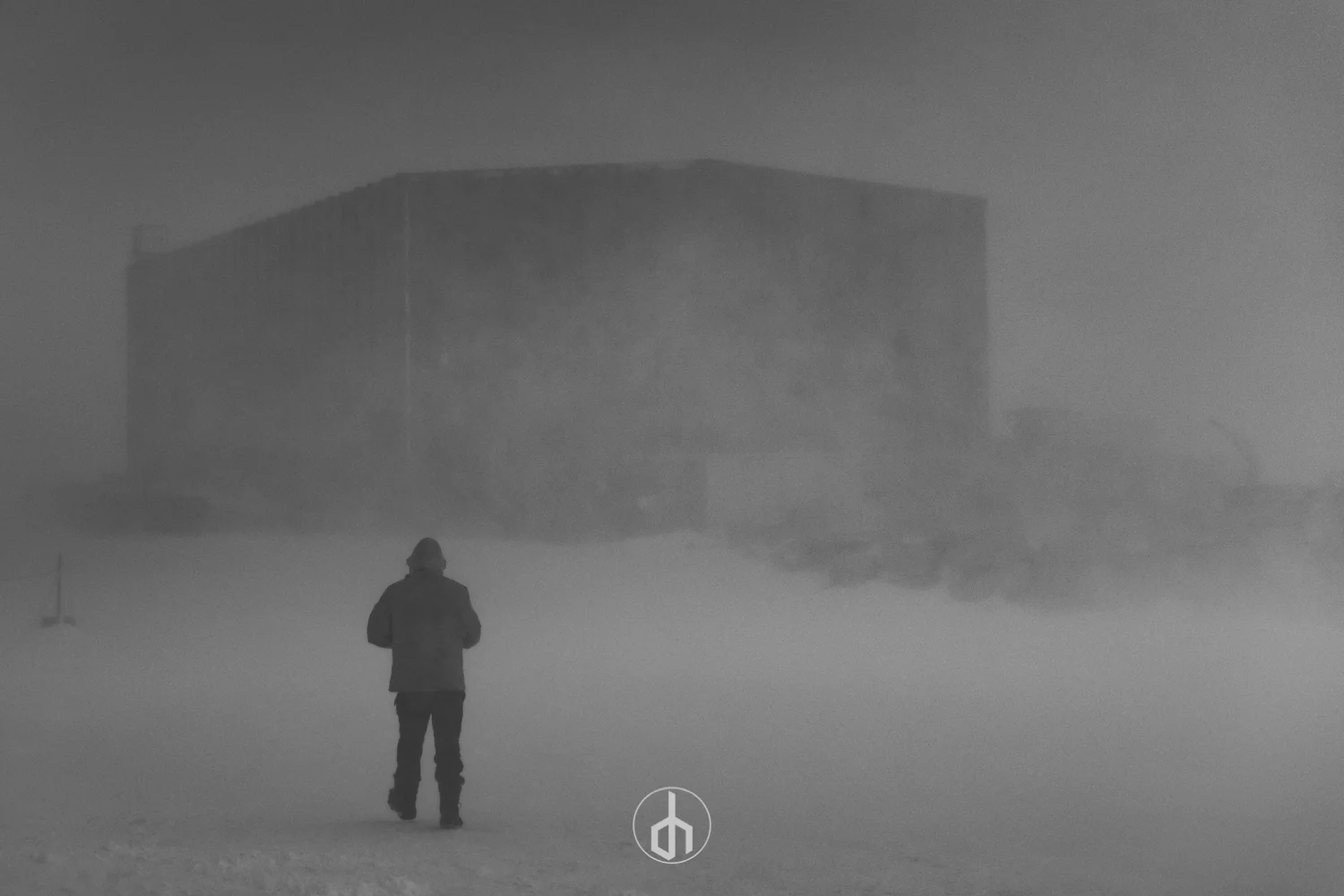 A person walking in a foggy, snow-covered landscape with a large, modern building in the background. There is a peace symbol at the bottom center of the image.
