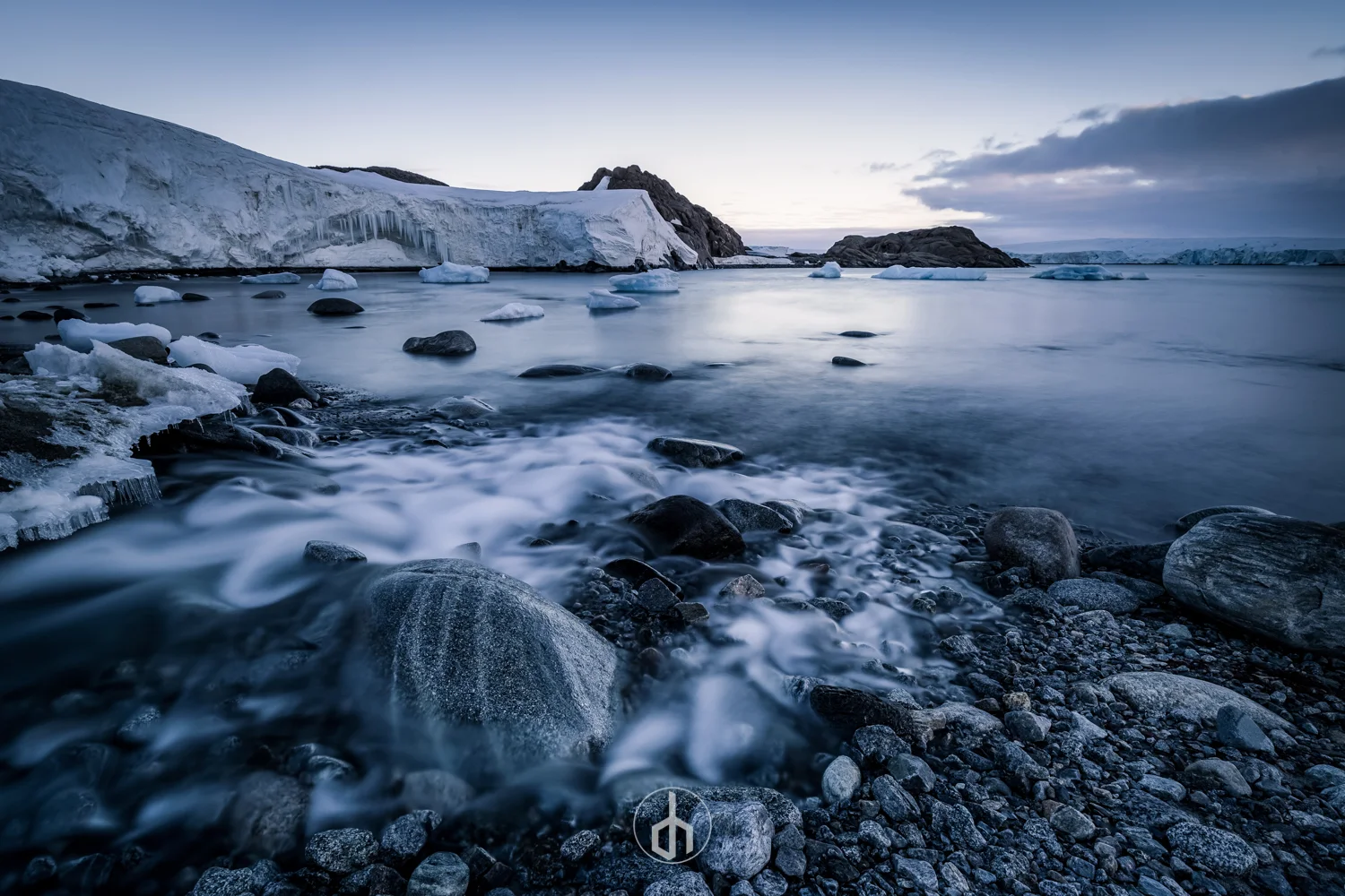 A cold, rocky shoreline with flowing water and floating ice under a cloudy sky, with ice-covered cliffs in the background.