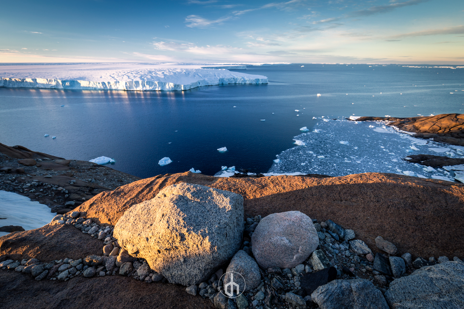 Arctic landscape featuring rocky terrain in the foreground, icy water with floating ice chunks, and a large iceberg in the distance under a partly cloudy sky.