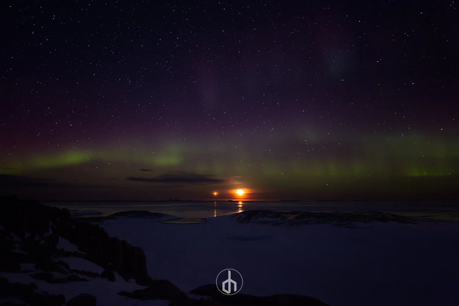 Nighttime landscape featuring the northern lights, stars, and a setting or rising moon over a snowy coastline.