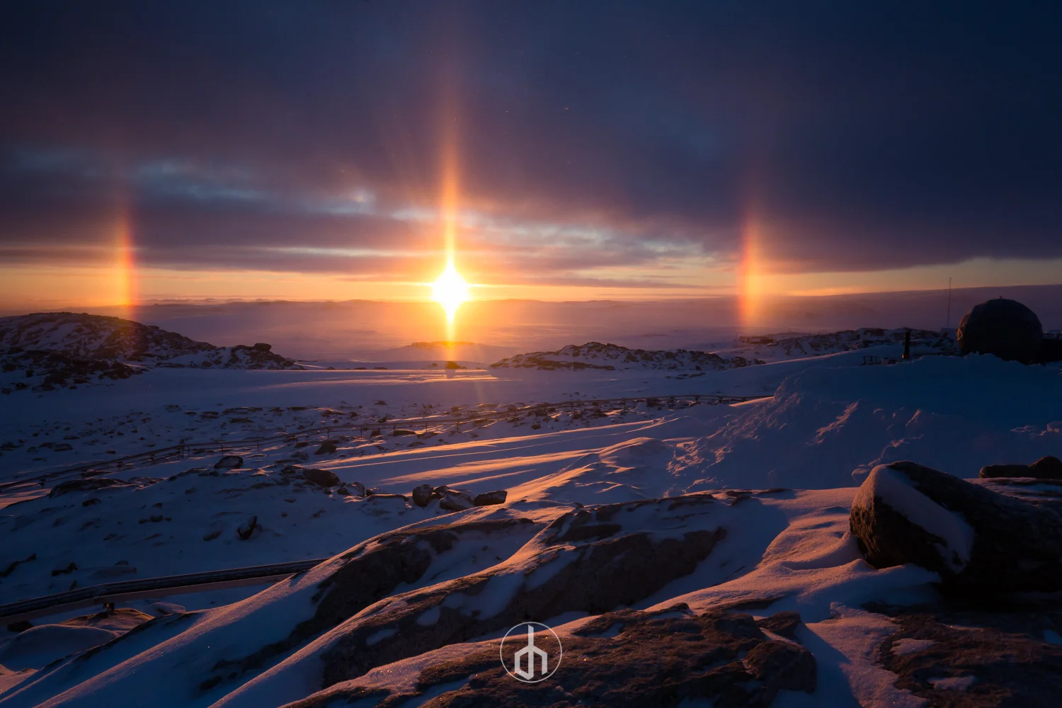 Snow-covered landscape at sunset with sun partly obscured by clouds, creating a sun halo and sundogs, with rocky terrain in the foreground.