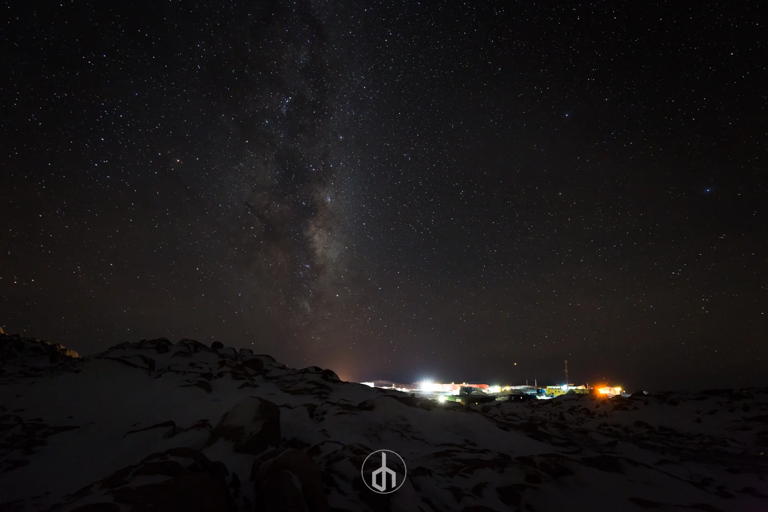 Night sky with stars and the Milky Way galaxy over a dark landscape with snow-covered ground and distant lights from a small town or settlement.