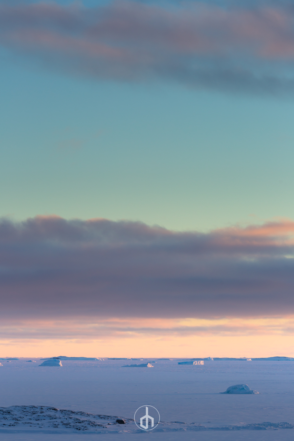 A polar landscape with ice-covered land, some icebergs in the water, and a sky with clouds during sunset or sunrise.
