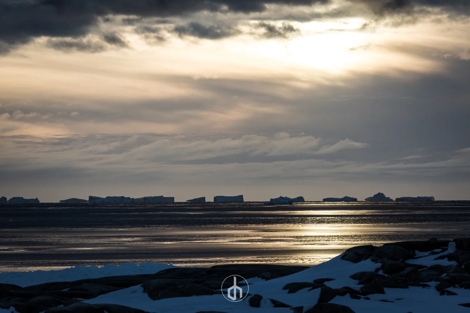 A landscape with icebergs floating on water under a cloudy sky during sunset or sunrise, with snow and rocks in the foreground.