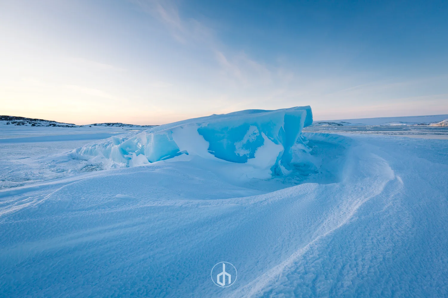 A large blue ice formation on a snow-covered landscape during sunset or sunrise, with a peace sign symbol at the bottom center of the image.